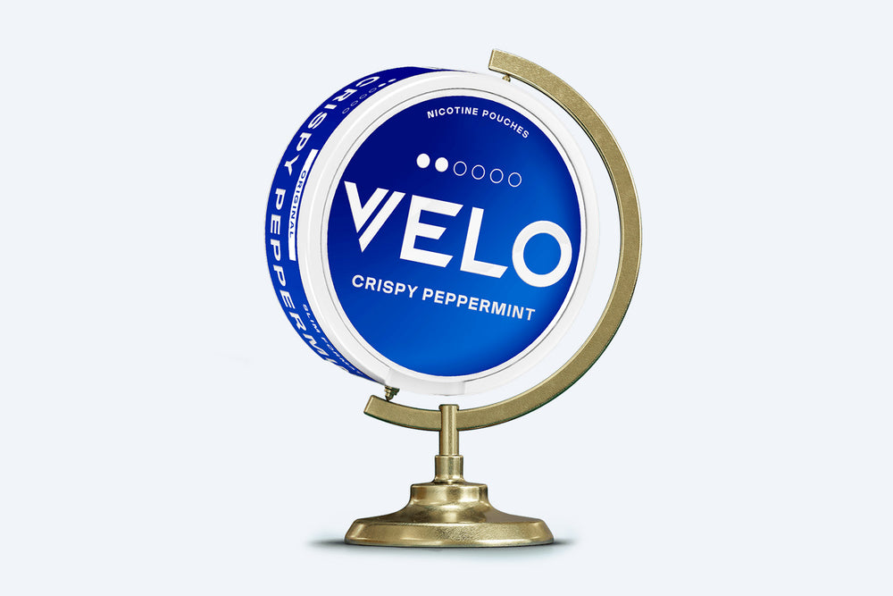 Velo Nicotine Pouches |  A round VELO Crispy Peppermint nicotine pouch container is displayed on a brass globe stand, with a blue and white label facing forward, highlighting the premium pouches inside.