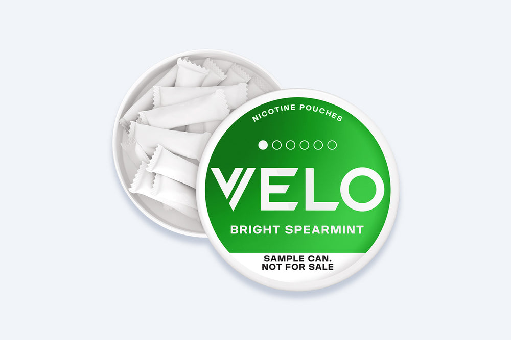 Velo Nicotine Pouches |  An open round can labeled “VELO Bright Spearmint” reveals white nicotine pouches inside. The lid states, “Sample can. Not for sale.” The green and white packaging prominently features the Velo branding, emphasizing these fresh spearmint nicotine pouches.