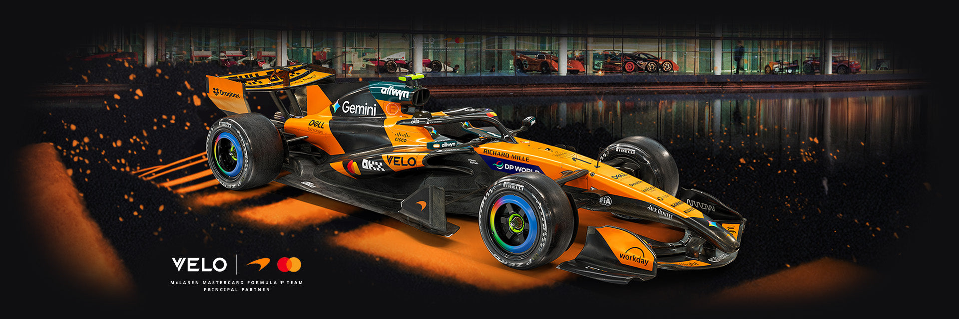 Velo Nicotine Pouches |  A bright orange and black Formula 1 car speeds forward, kicking up orange sparks. The background shows a garage with cars. Logos such as VELO—known for their nicotine pouches—and Gemini are visible on the car.