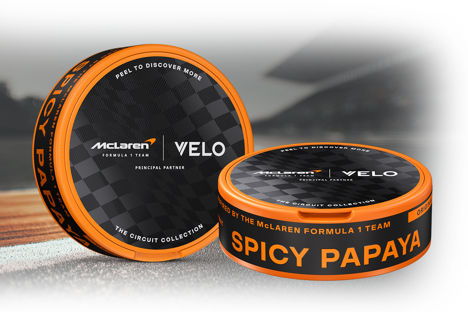 Velo Nicotine Pouches |  Two circular tins with orange rims labeled “VELO” and “SPICY PAPAYA,” featuring McLaren Formula 1 branding, sit atop a racetrack-themed background—each tin filled with Velo nicotine pouches for a sleek, invigorating experience.