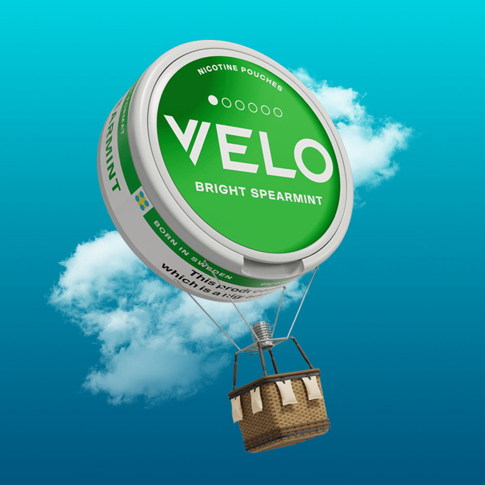Velo Nicotine Pouches |  A green VELO nicotine pouch tin floats in the sky like a hot air balloon, with a wicker basket attached underneath, carrying pouches against a blue sky with clouds.