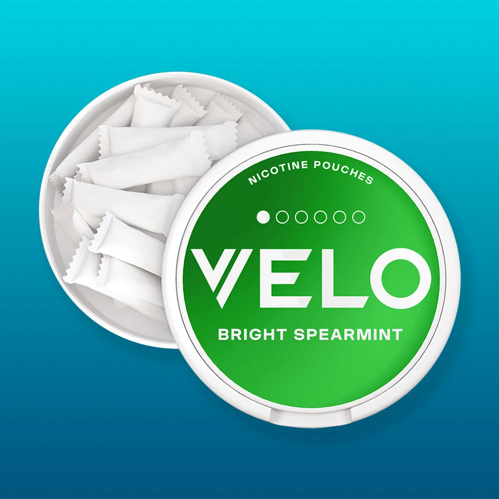 Velo Nicotine Pouches |  A round container of VELO Bright Spearmint nicotine pouches, partially open to show the white pouches inside. The VELO-branded lid is green with white text and sits on a blue gradient background.