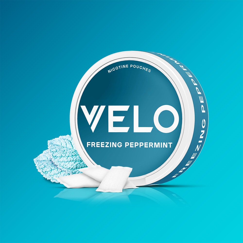 Velo Nicotine Pouches |  A round container labeled VELO Freezing Peppermint is shown with white nicotine pouches—some opened to reveal blue peppermint powder—on a blue gradient background, embodying the intensely strong menthol sensation of VELO’s Freezing Peppermint.