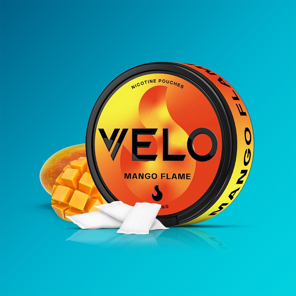 Velo Nicotine Pouches |  A round orange VELO Mango Flame nicotine pouch container features a flame motif, sliced mango imagery, and white pouches on a blue background. The container prominently displays the words “VELO” and “Mango Flame,” emphasizing its heated mango flavour for an intense burning sensation.