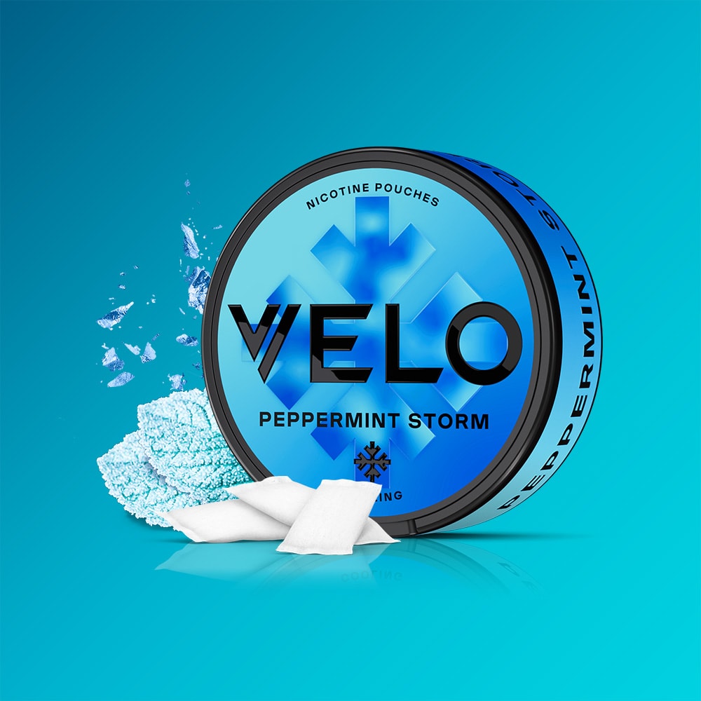 Velo Nicotine Pouches |  A round blue tin of VELO Peppermint Storm rests on a blue background, accompanied by white nicotine pouches and scattered crushed peppermint. The tin displays a frosty, snowflake-inspired design that reflects the intense blend of peppermint and menthol inside, offering an invigorating cooling sensation.