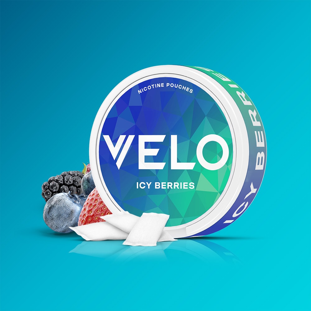 Velo Nicotine Pouches |  A round container of VELO Icy Berries sits on a blue background, accompanied by fresh berries and two white nicotine pouches, showcasing the triple-berry blend and refreshing cooling sensation of each pouch.
