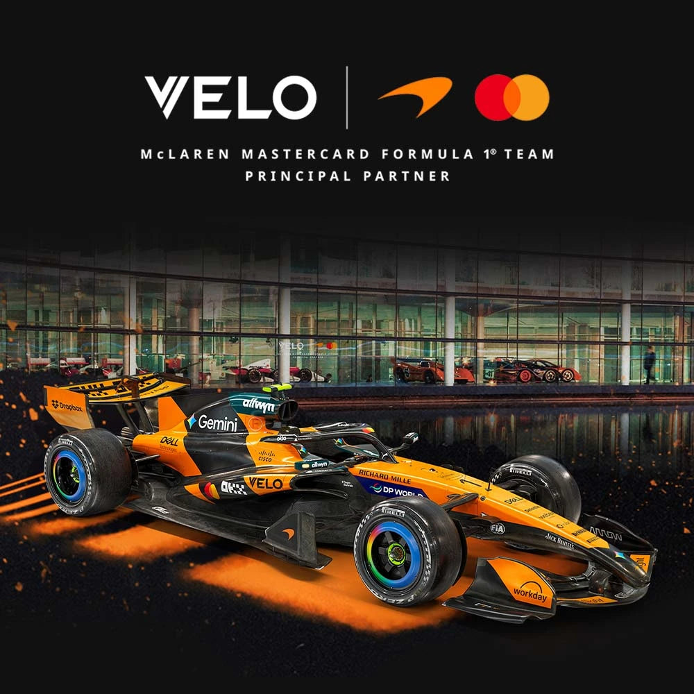 Velo Nicotine Pouches |  An orange and black McLaren Formula 1 car is shown in front of a glass-walled garage. Above it are the Velo, McLaren, and Mastercard logos, along with the text McLaren Mastercard Formula 1 Team Principal Partner. The prominent Velo logo highlights their nicotine pouches partnership with the team.