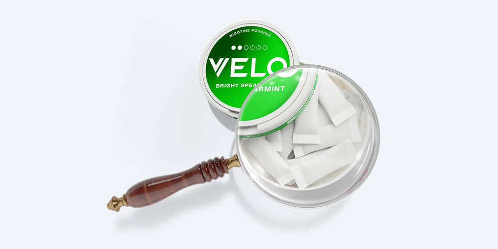 Velo Nicotine Pouches |  A round tin labeled VELO Bright Spearmint is displayed, featuring sleek white nicotine pouches, with a magnifying glass highlighting the Velo pouches for a closer look.