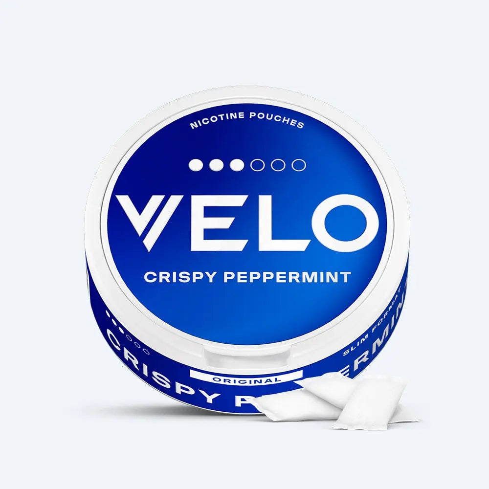 Velo Nicotine Pouches |  A round, blue and white container labeled VELO Crispy Peppermint sits prominently, showcasing the Velo nicotine pouches inside, with two white pouches placed neatly in front of it on a light background.