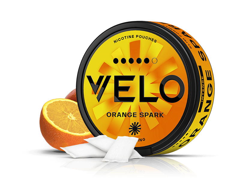 Velo Nicotine Pouches |  Round VELO Orange Spark nicotine pouch container featuring a vibrant orange and yellow design. Two white pouches are displayed in front alongside a sliced orange, all set against a white background—conjuring the fresh aroma of ripe oranges for a truly citrus-inspired experience.
