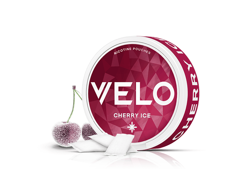 Velo Nicotine Pouches |  A round container of VELO Cherry Ice nicotine pouches stands next to two frosty cherries and several Velo pouches, all on a white background.