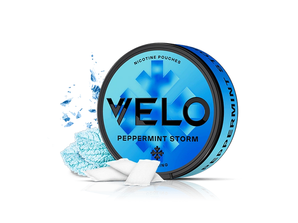Velo Nicotine Pouches |  A blue tin of VELO Peppermint Storm nicotine pouches sits on a white background, with several Velo pouches and a sprinkle of blue powder arranged artfully beside it.