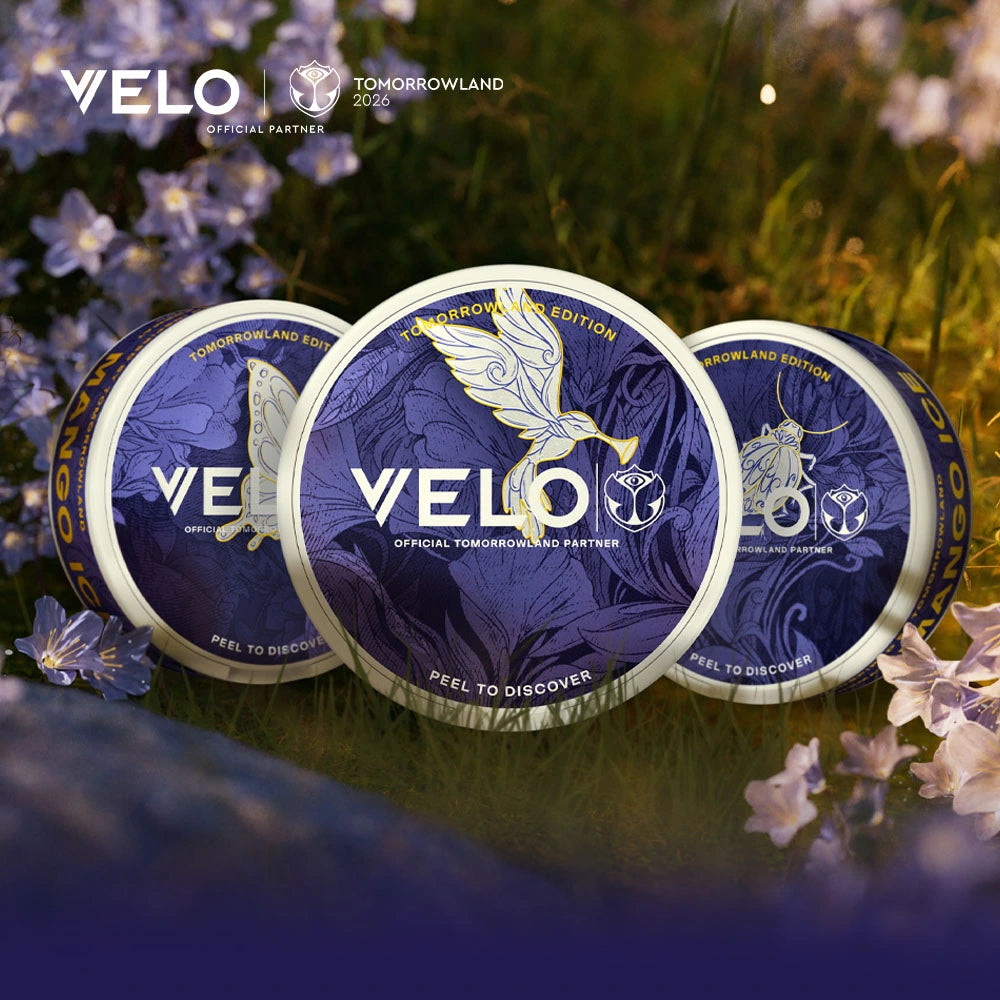 Velo Nicotine Pouches |  Three VELO nicotine pouches in purple “Tomorrowland Edition” packaging sit on grass with small white flowers. A gold bird and Tomorrowland logo appear on the VELO containers, emphasizing their unique design. The background has a soft, dreamy look.